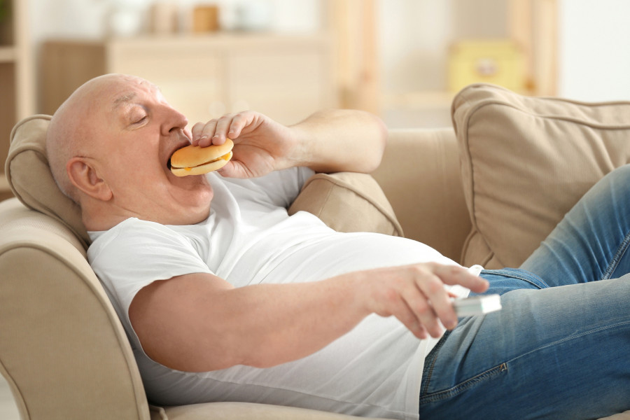 man watching TV and eating hamburger while lying on sofa couch. Sedentary lifestyle